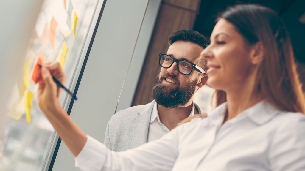 Kobieta i mężczyzna stoją przed tablicą. Kobieta pisze po przyczepionej do niej kartce.